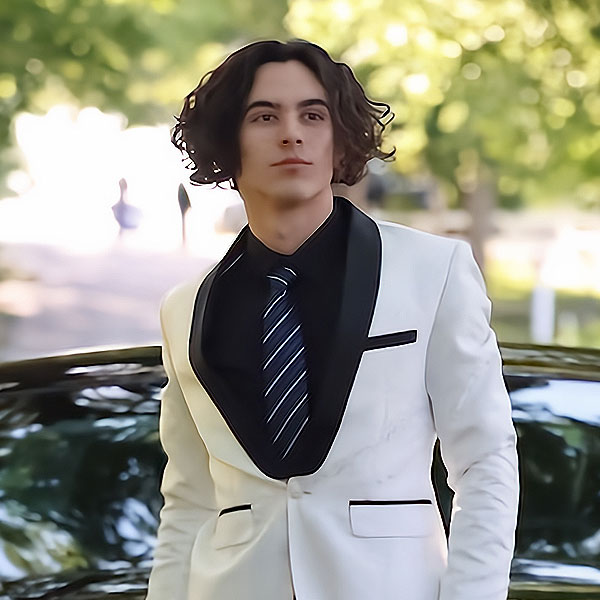 Senior Spotlight: A young man with curly hair wears a white tuxedo jacket, black shirt, and striped tie, standing outdoors in front of a car with trees and bright sunlight—celebrating the Class of 2023.