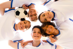 A group of five smiling children, looking like a joyful team at Spare Time Texas, huddle together in white soccer uniforms with blue trim, viewed from below. One child holds a soccer ball as they exhibit the united spirit often found at a family fun center.
