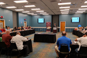 In a vibrant conference room with blue walls and a carpeted floor, people are seated around a U-shaped table setup. A speaker presents at a podium in front of two screens. It’s reminiscent of the engaging atmosphere found at Spare Time Texas, offering inspiration beyond just bowling and family fun.