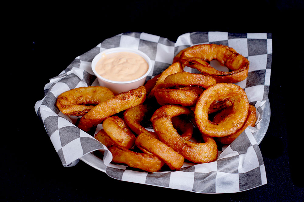 A basket of golden-brown onion rings on a black and white checkered paper, served with a small cup of dipping sauce, sits invitingly at Spare Time Texas. Perfect for sharing at this lively family fun center where bowling is just the beginning of the excitement.