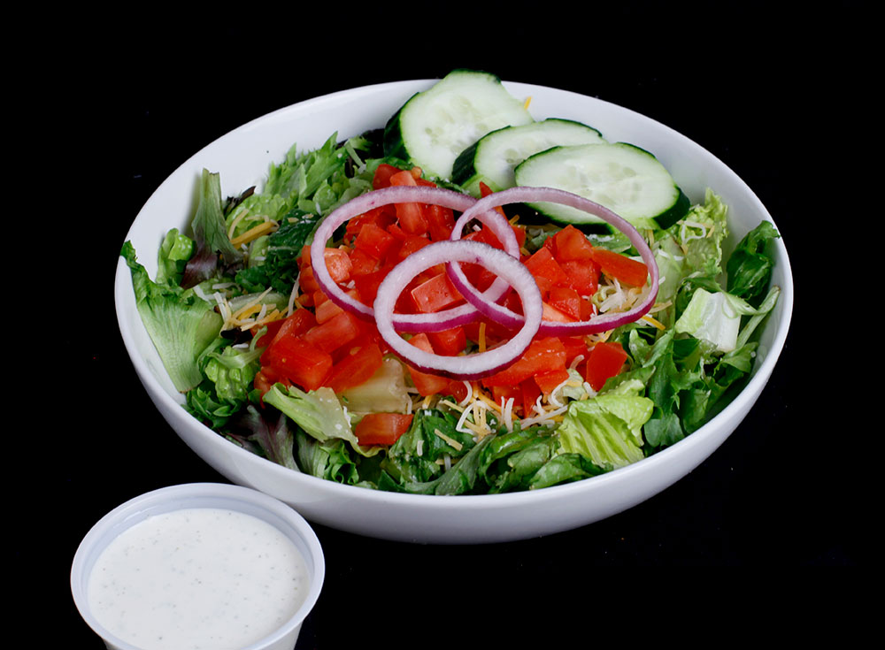 At Spare Time Texas, enjoy a fresh salad with sliced cucumbers, tomatoes, red onions, and mixed greens in a white bowl. It's the perfect bite as you take a break from the excitement of bowling or sharing laughs at our family fun center. A small container of creamy dressing is served on the side.