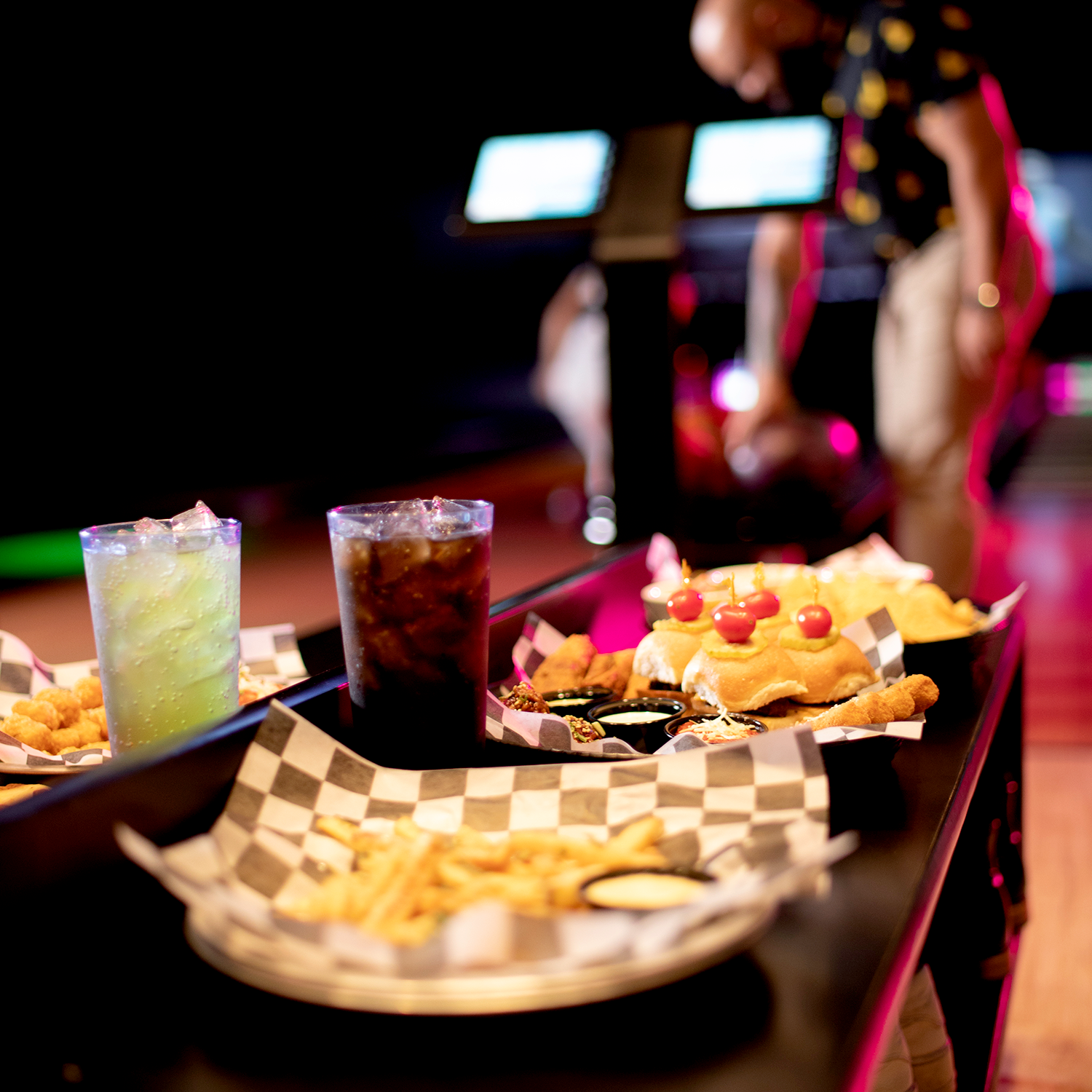 A close-up of a bowling alley table at Spare Time Texas features a hamburger topped with cherry tomatoes, fries, chicken tenders, and two drinks. In the background, a person is seen preparing to bowl at this family fun center.