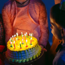 In the dimly lit room at Spare Time Texas, a child gazes at a birthday cake adorned with flickering candles. The yellow-frosted delight is held by someone in a pink top, capturing the essence of joy in this special celebration moment.