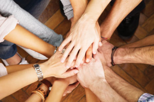 A close-up of multiple people's hands stacked on top of each other, symbolizing unity and teamwork at Spare Time Texas. The diverse skin tones against the wooden floor reflect the vibrant community spirit in this family fun center.