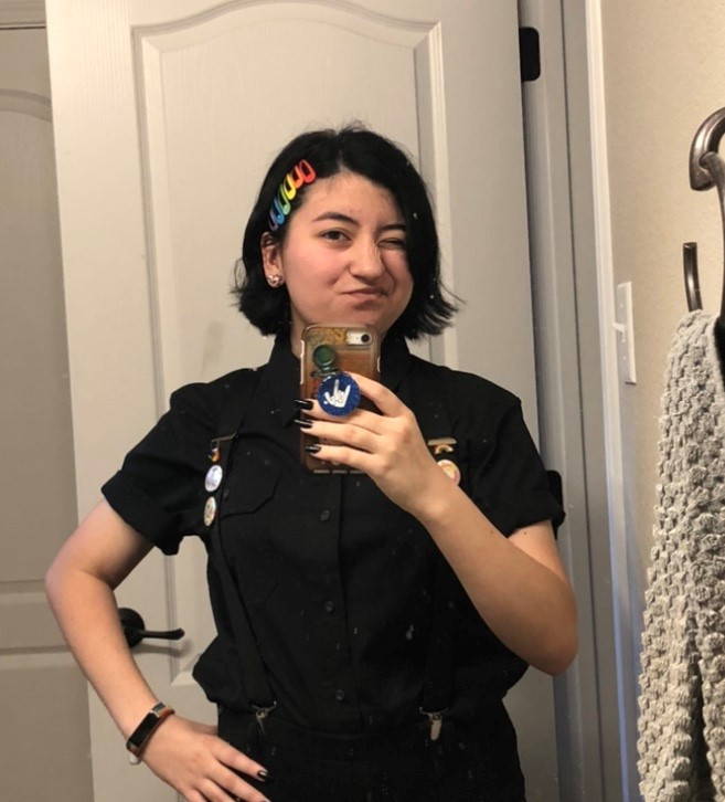 Person with black hair and colorful hair clips takes a mirror selfie at Spare Time Texas. They’re wearing a black shirt with buttons and suspenders. A towel hangs on a nearby rack. Winking, they hold a phone adorned with a blue sticker, perhaps capturing memories in this family fun center.