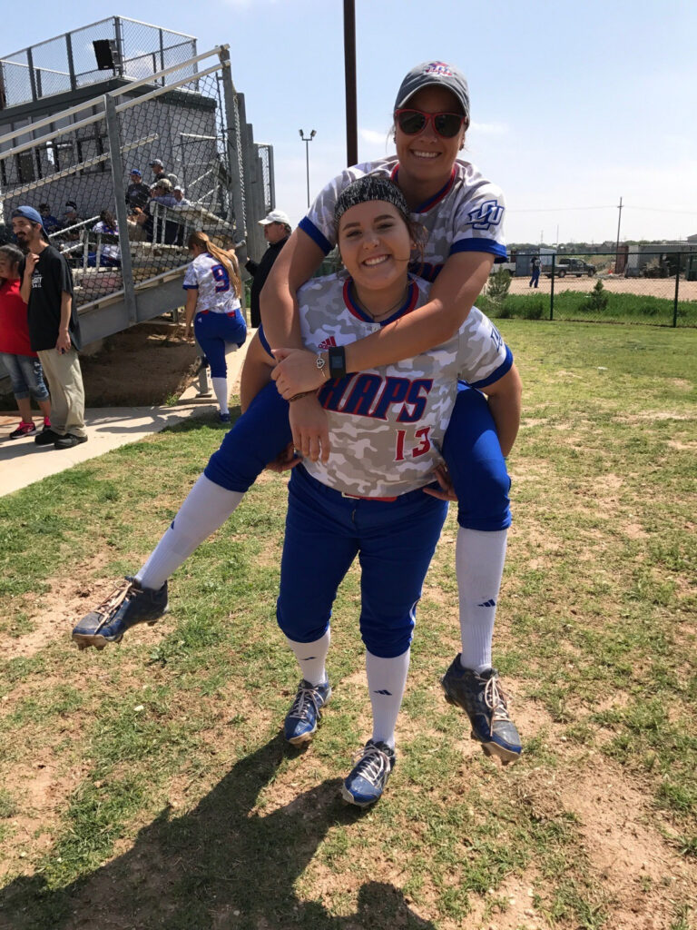 Two softball players in blue and white uniforms enjoy a playful piggyback ride on the grassy field, reminiscent of the joy found at Spare Time Texas. They are smiling, with bleachers and a few people in the background on this sunny day.