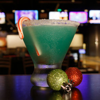 A blue cocktail in a glass with a sugared rim is garnished with a candy cane, perfect for enjoying at Spare Time Texas. Two glittery Christmas ornaments, one green and one red, add festivity as blurred TV screens enhance the lively atmosphere of this family fun center.