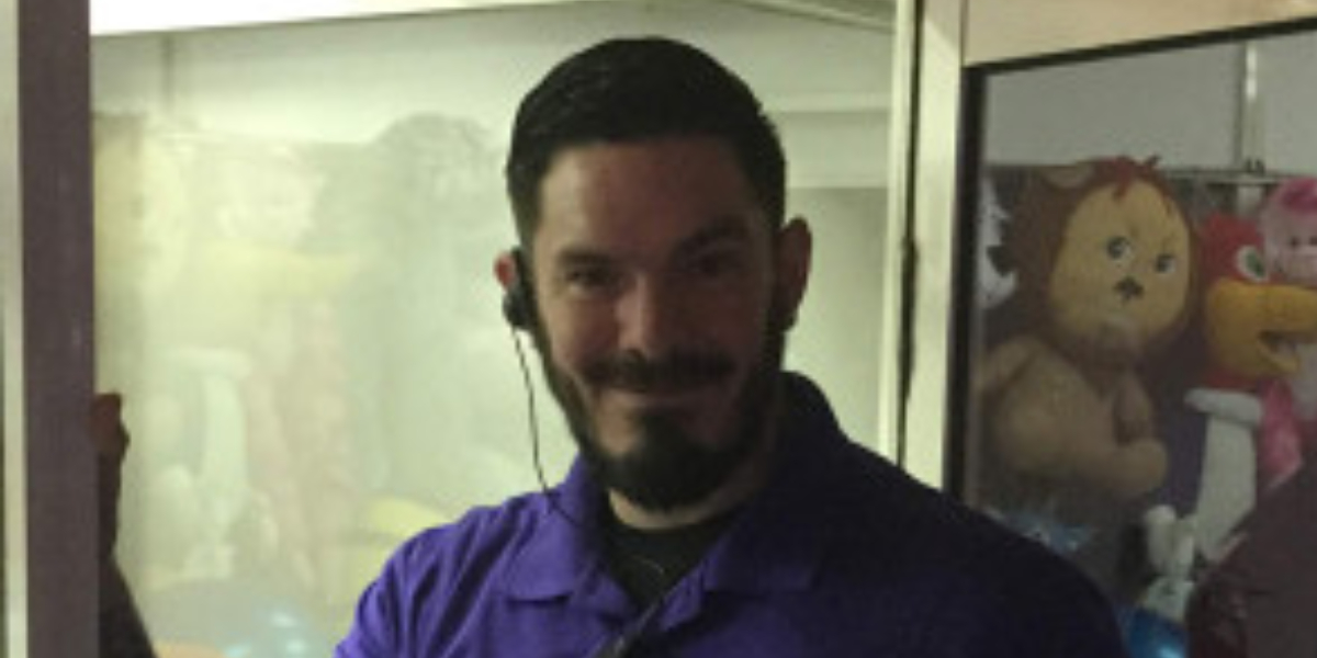 A person with a beard, wearing a purple shirt and earpiece, stands smiling at the camera in a room at Spare Time Texas. Through the glass window behind them, plush toys hint at the family fun center's playful atmosphere.