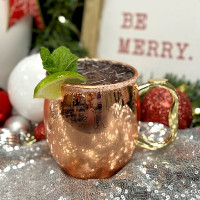 A copper mug filled with a dark beverage, garnished with a lime wedge and mint sprig, sits beside red and white ornaments. Greenery adds cheer to the scene, while a "Be Merry" sign captures the spirit—perfect for enjoying at Spare Time Texas during some festive bowling fun.