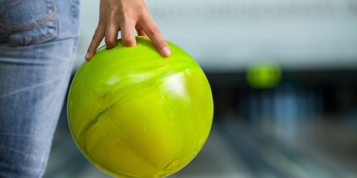 Close-up of a person's hand holding a bright green bowling ball at Spare Time Texas, preparing to bowl. The background shows blurred lanes and pins, highlighting the focus on the ball and hand. The person is wearing jeans, ready for action in this family fun center.