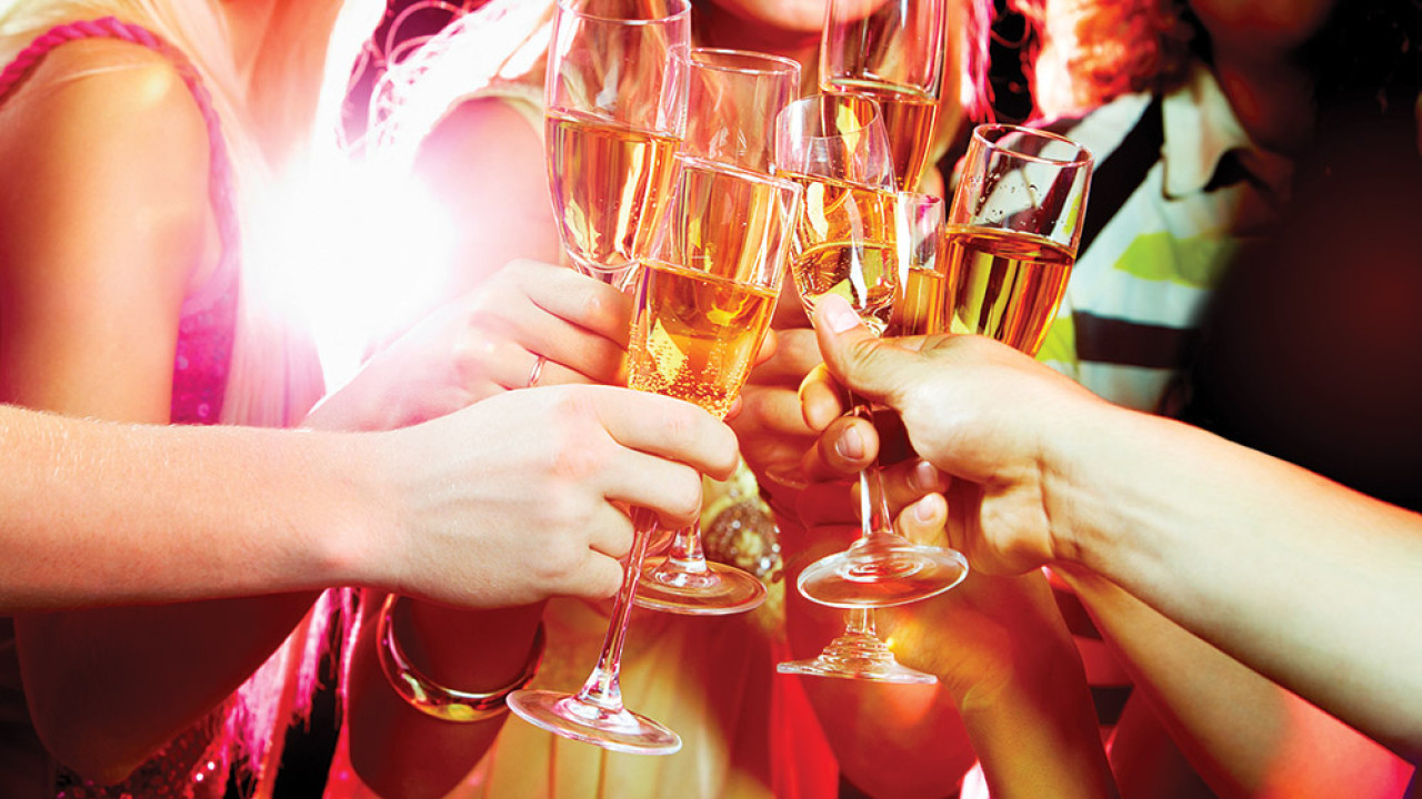 A group of people raising champagne glasses in a toast at Spare Time Texas, bathed in warm lighting. The image captures a celebratory moment with blurred figures from the family fun center in the background and a bright light source on the left.