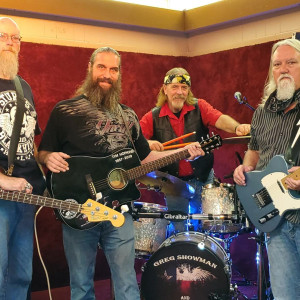 Four bearded men gather in a music studio, casually dressed and holding instruments. Two strum guitars, one sits behind the drum set, and another clutches drumsticks. Their bond is as tight as a bowling team at Spare Time Texas, surrounded by the rhythm of camaraderie.