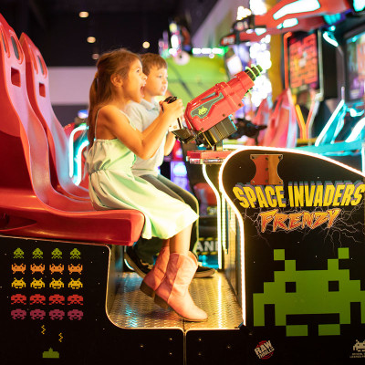 Two children in colorful clothes play an arcade game called "Space Invaders Frenzy" at Spare Time Texas. Seated on red chairs, they aim with joysticks as bright neon lights illuminate the family fun center, creating a vibrant atmosphere perfect for bowling and endless entertainment.