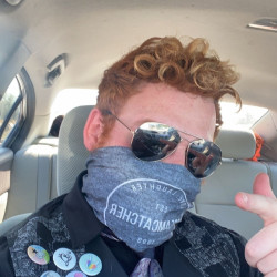 In their spare time, this person with curly hair and sunglasses enjoys hitting the bowling lanes at a local family fun center in Texas. Now, they're sitting stylishly in a car, sporting a patterned face covering and a jacket decked out with pins while making an animated hand gesture.