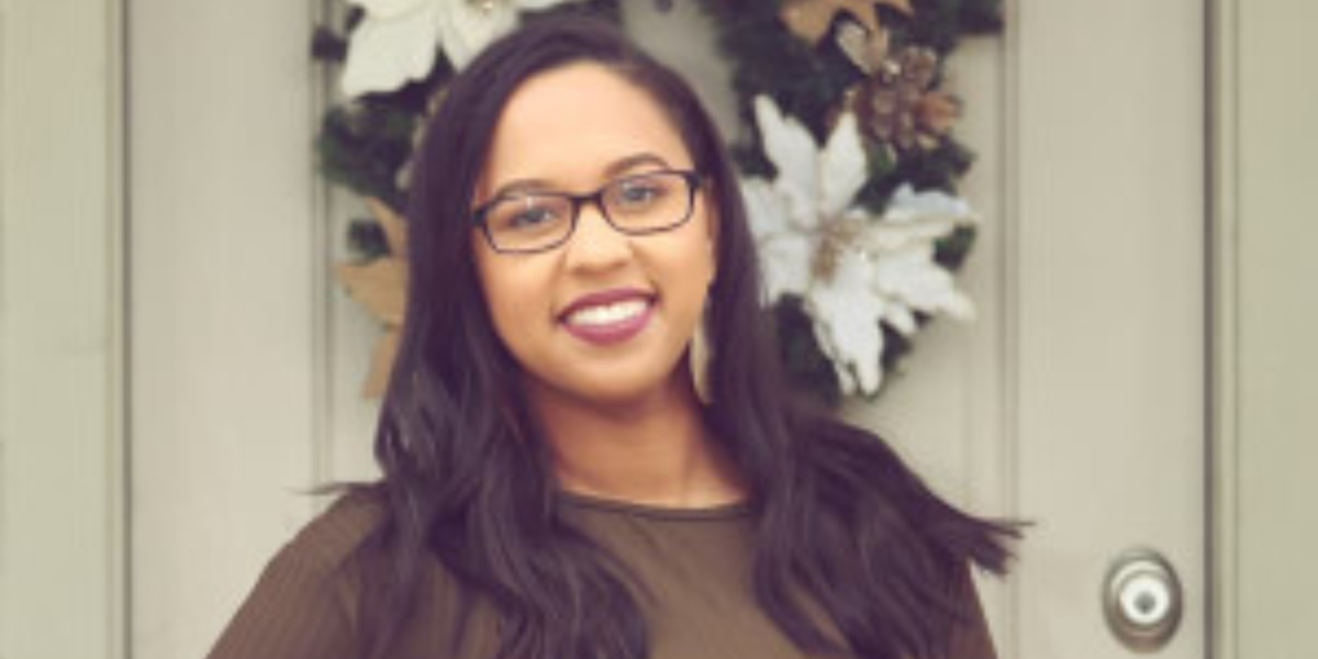 A person with long dark hair and glasses smiles while standing in front of a door decorated with a festive wreath featuring white and gold elements, ready for an exciting day at Spare Time Texas, where family fun and bowling await.