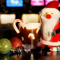 A festive scene at Spare Time Texas features a small Santa figurine next to a mug of hot chocolate topped with a candy cane. Three glittery ornaments—red and green—glow nearby, set against a blurred background of colorful lights, capturing the spirit of bowling and family fun.
