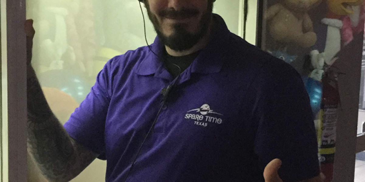 A man in a purple "Spare Time Texas" shirt stands inside a phone booth, smiling and giving a thumbs up. He has a beard and tattoos on his arm. The vibrant background hints at the lively atmosphere of the family fun center, complete with colorful objects and a fire extinguisher.