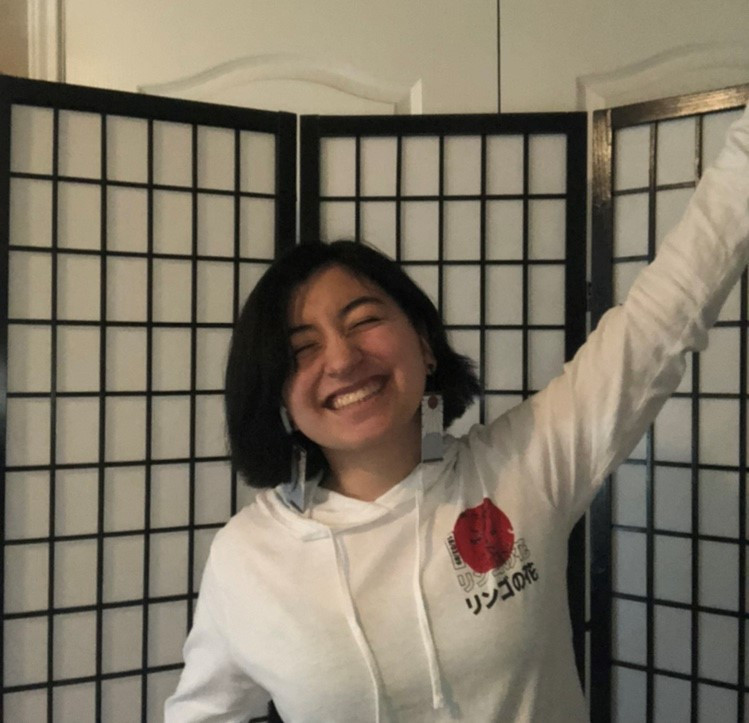 A person with short dark hair is smiling broadly, raising one arm in front of a shoji screen. They're sporting a white hoodie featuring a red rose and Japanese characters, looking ready for a thrilling day of bowling at Spare Time Texas.