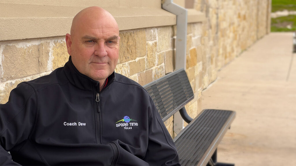 A person wearing a black jacket with "Coach Dave" and a logo sits on a bench against a stone wall at the family fun center. The background includes a walkway and grass, hinting at the lively atmosphere of Spare Time Texas. The weather appears cloudy.