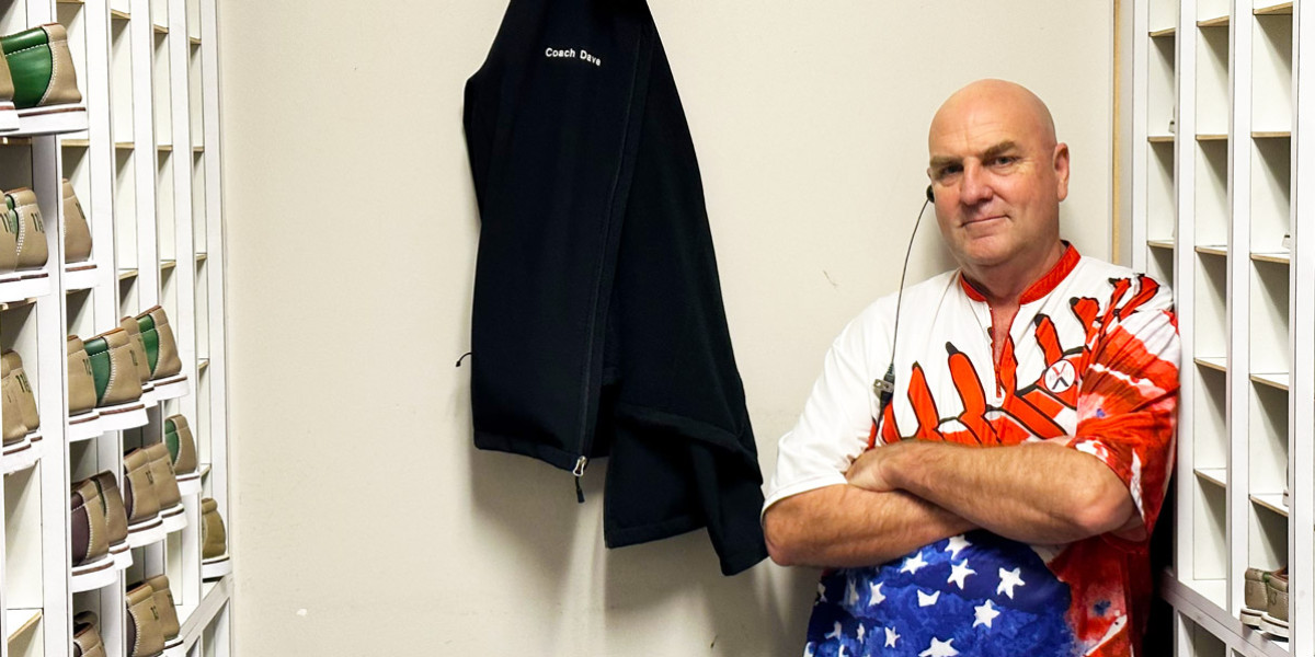 A man in a brightly colored shirt stands with arms crossed in a room filled with rows of shelves holding beige hats, reminiscent of the vibrant vibe at Spare Time Texas. A black jacket with "Coach Dug" written on it hangs on the wall beside him, echoing the fun-loving spirit found at a family fun center.