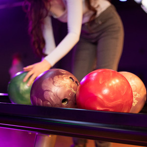 In the dimly lit bowling alley of Spare Time Texas, a person reaches for a bowling ball from the rack. The rack holds four colorful balls—green, purple, red, and pink—awaiting their turn to roll down the lane in this lively family fun center.