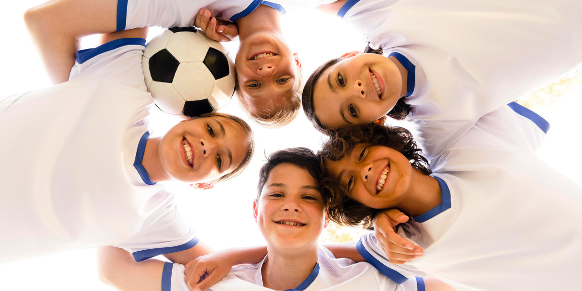 Five children in white soccer uniforms with blue trim form a huddle, smiling up at the camera. One holds a soccer ball as they enjoy their day outdoors under a bright sky, reminiscent of the camaraderie you'd find at Spare Time Texas.