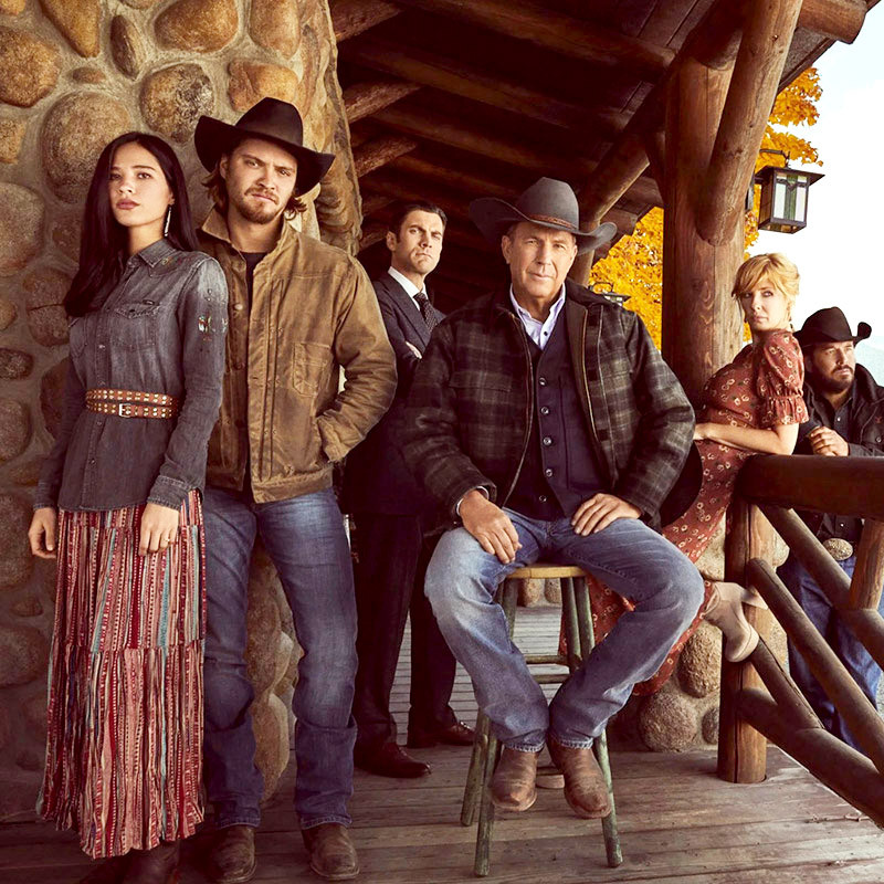 A group of people in Western attire pose on a wooden porch at Spare Time Texas. Some wear cowboy hats and jeans, while others don dresses. They stand or sit against a rustic stone and wood backdrop, with autumn leaves visible in the background, capturing the essence of family fun.
