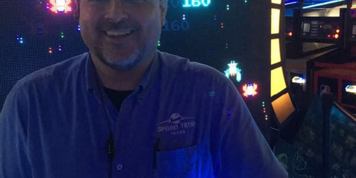 In the dimly lit confines of a family fun center, a person smiles in front of a vintage arcade game machine with colorful screens. Wearing a light blue shirt with an embroidered logo and badge, they revel in the retro gaming ambiance at Spare Time Texas.