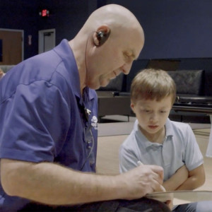 A bald man in a blue shirt and earpiece writes on a clipboard beside a young boy in a light blue shirt at Spare Time Texas. Indulging in family fun, the pair sits indoors, rows of chairs around them, as the boy looks on attentively amidst the lively chatter of the family fun center.