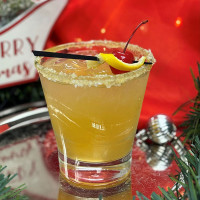 A festive cocktail in a glass with a sugared rim, garnished with a cherry and lemon twist skewer. The background features a "Merry Christmas" sign and red fabric reminiscent of holiday gatherings at Spare Time Texas, where family fun abounds amid the green foliage and lively bowling lanes.