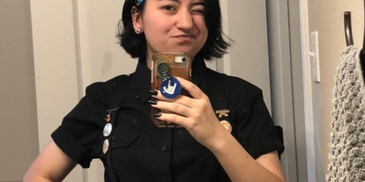 A person with short black hair, wearing a black shirt and blue headband, takes a selfie in a mirror. They're holding a phone with a unique case and have a playful expression, perhaps after enjoying some bowling at their favorite family fun center. Various pins are attached to their shirt.