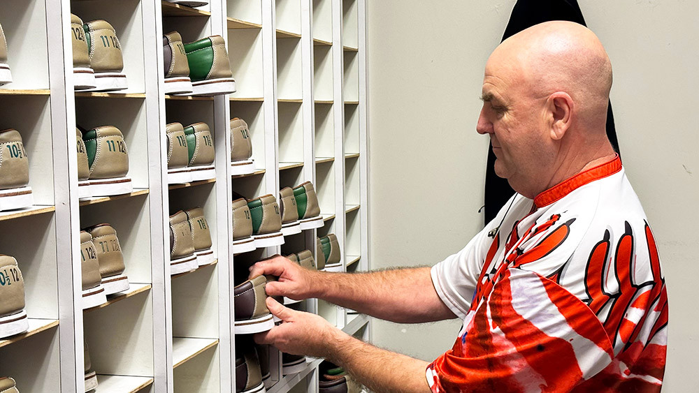 In his spare time, a man in a red and white shirt organizes a collection of beige and green shoes in a wall-mounted cubby shelf at the family fun center. Each cubby holds a pair with numbers on the heels as he carefully places them in their spots, ensuring everything is ready for the next round of bowling.