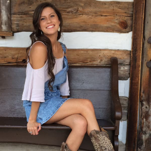 A woman with long hair sits on a wooden bench in front of a rustic log wall at Spare Time Texas. She wears a light blouse, denim overalls, and leopard print boots, smiling at the camera with her legs crossed.