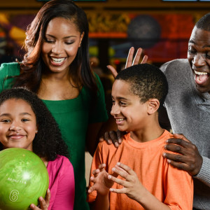 A happy family of four enjoying a bowling outing at Spare Time Texas. The young girl holds a green bowling ball, while everyone smiles and looks at each other with excitement. They appear to be having a joyful and fun time together at this vibrant family fun center.