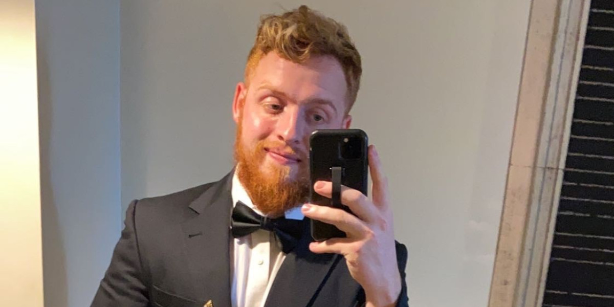 A person with a reddish beard takes a mirror selfie, wearing a dark suit with a bow tie and smiling slightly, as if ready for an evening out at Spare Time Texas. The background features a light wall and a partial view of a doorway.
