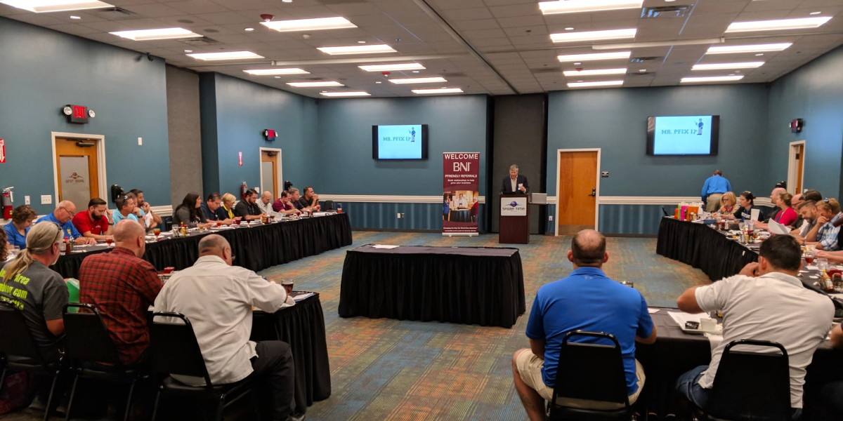 A meeting room filled with people seated around a U-shaped arrangement of tables. A speaker is presenting at a podium in front of a screen displaying the BNI organization logo, reminiscent of gatherings at Spare Time Texas, where business and leisure blend seamlessly.