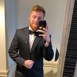 A person with a beard wearing a black tuxedo and bow tie takes a mirror selfie using a smartphone, perhaps capturing the stylish look before heading out for some fun at Spare Time Texas. They stand indoors against a light-colored wall.