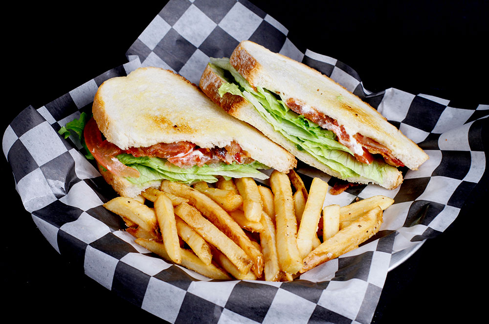 A sandwich cut in half, filled with lettuce, tomato, bacon, and mayonnaise, is placed on checkered paper. It's the perfect meal at Spare Time Texas after a game of bowling or enjoying the arcade at this family fun center. Served alongside a portion of golden brown French fries.