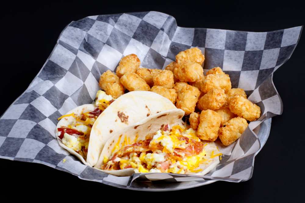 Two breakfast tacos with scrambled eggs, bacon, and cheese are served on a checkered paper-lined plate alongside crispy tater tots, making the perfect start to a day of family fun at Spare Time Texas.