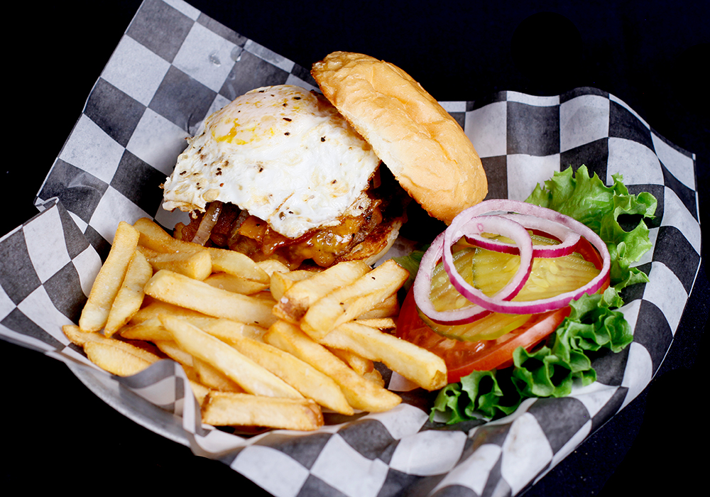 Indulge in a Spare Time Texas special: a burger with a fried egg, bacon, and cheese in a bun, served with crispy fries. This family fun center favorite includes lettuce, tomato, pickles, and red onion on classic checkered paper. Perfect after an exciting round of bowling!.