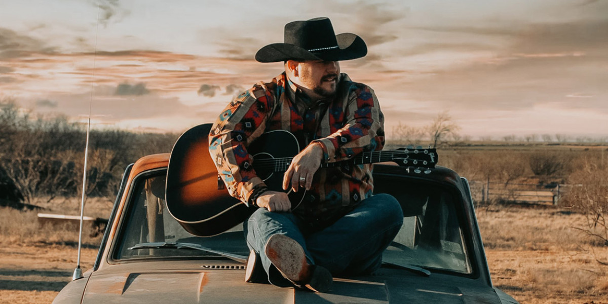 Jon Hope. A person wearing a cowboy hat and patterned jacket sits on an old truck's hood, holding a guitar. The setting is a rural landscape with a cloudy sky at sunset, reminiscent of the scenic backdrop you'd find at Spare Time Texas after enjoying some family fun.