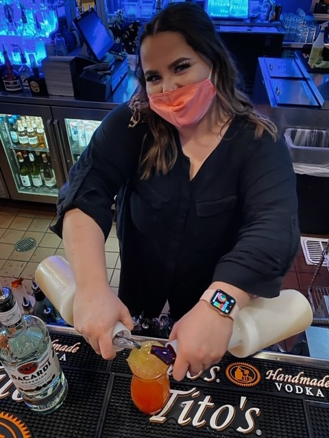 A bartender wearing a pink face mask and black shirt is artfully pouring drinks using two bottles at Spare Time Texas. She stands behind the bar counter, smiling amid various liquors and bar tools, while her Apple Watch glints under the lights of this lively family fun center.