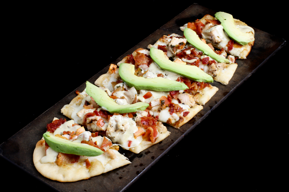 A rectangular flatbread topped with melted cheese, grilled chicken, tomato pieces, and bacon bits is perfect for your next visit to Spare Time Texas. Fresh avocado slices are neatly arranged on top, served on a dark serving platter—ideal for sharing after a game of bowling.