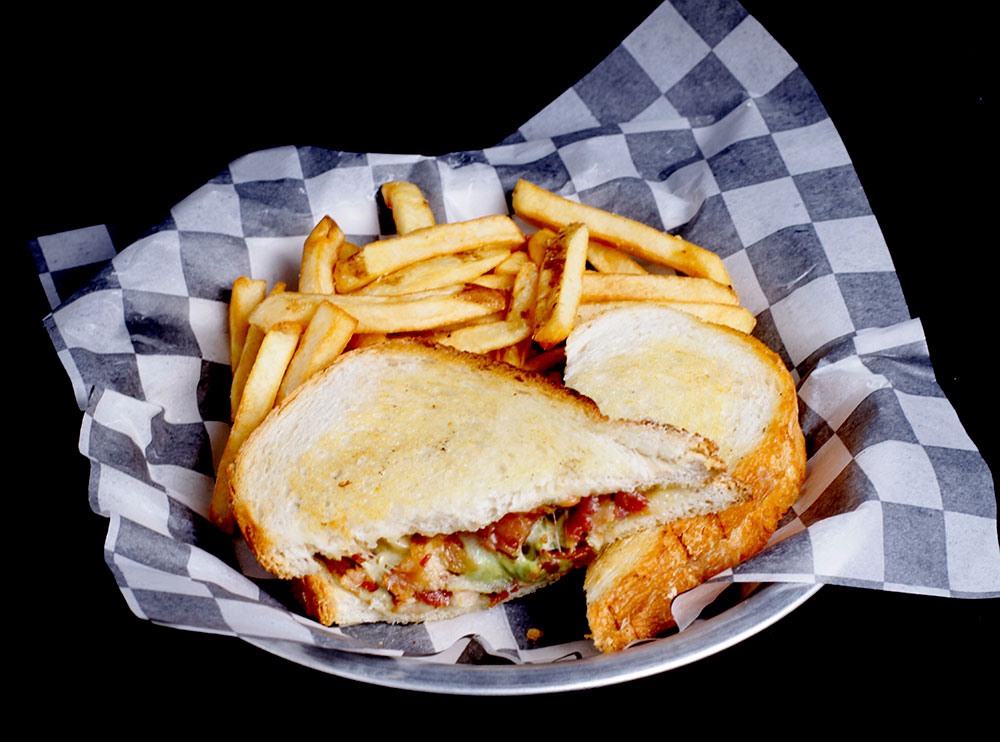 At Spare Time Texas, indulge in a grilled sandwich with melted cheese and bacon, served on checkered paper alongside golden French fries. The black background highlights this delicious meal, perfect for enjoying after a lively game of bowling at our family fun center.