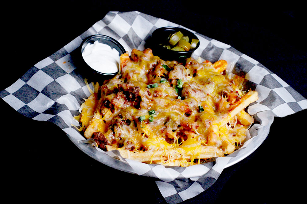 A plate of loaded cheese fries, perfect for sharing after a round of bowling at Spare Time Texas, topped with chopped green onions and crispy bacon on classic black and white checkered paper. Served with sides of sour cream and pickles in small black cups for that extra tangy kick.