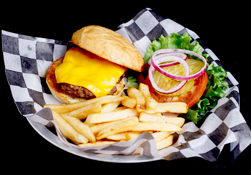 A cheeseburger with lettuce, tomato, pickles, and onions is served on checkered paper with a side of French fries at Spare Time Texas. The burger sits on a bun with melted cheese, perfect fuel after hitting the lanes for some family fun bowling.