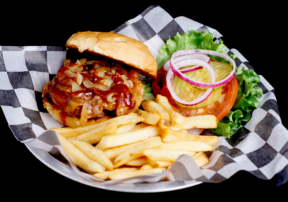 At Spare Time Texas, enjoy a cheeseburger topped with onions and barbecue sauce, nestled on a checkered paper-lined plate beside crispy french fries. Garnishes of lettuce, tomato, pickles, and red onion slices complete your meal at this family fun center where bowling is just the beginning of the excitement.