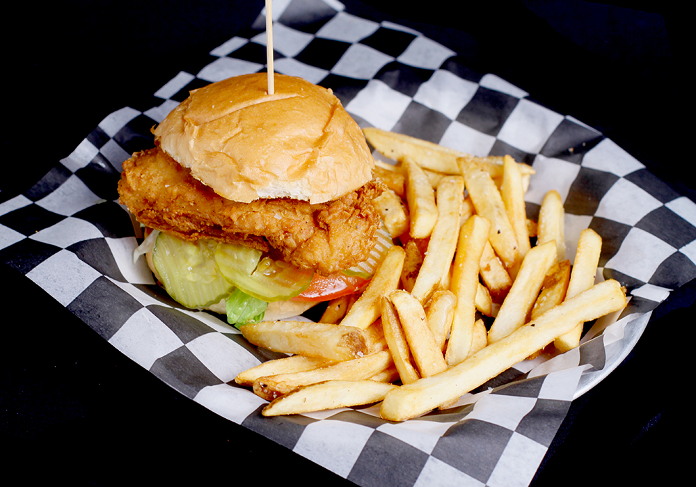 Enjoy a crispy fried chicken sandwich with lettuce, tomato, and pickles on a bun, served on a black and white checkered paper tray at Spare Time Texas. Accompanied by golden French fries, it’s the perfect treat after an exciting game of bowling at this family fun center.