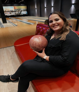 A person at Spare Time Texas is sitting on a red couch, smiling while holding a bowling ball. The lanes and pins are visible in the dimly lit background of this bustling family fun center, making for an enjoyable and entertaining atmosphere.