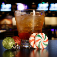 A glass of iced brown cocktail sits beside two green and red glittery ornaments and two round peppermint candies, capturing the festive vibe at Spare Time Texas. The dark, blurred background with hints of colorful lights echoes the lively atmosphere you'd find in a family fun center.
